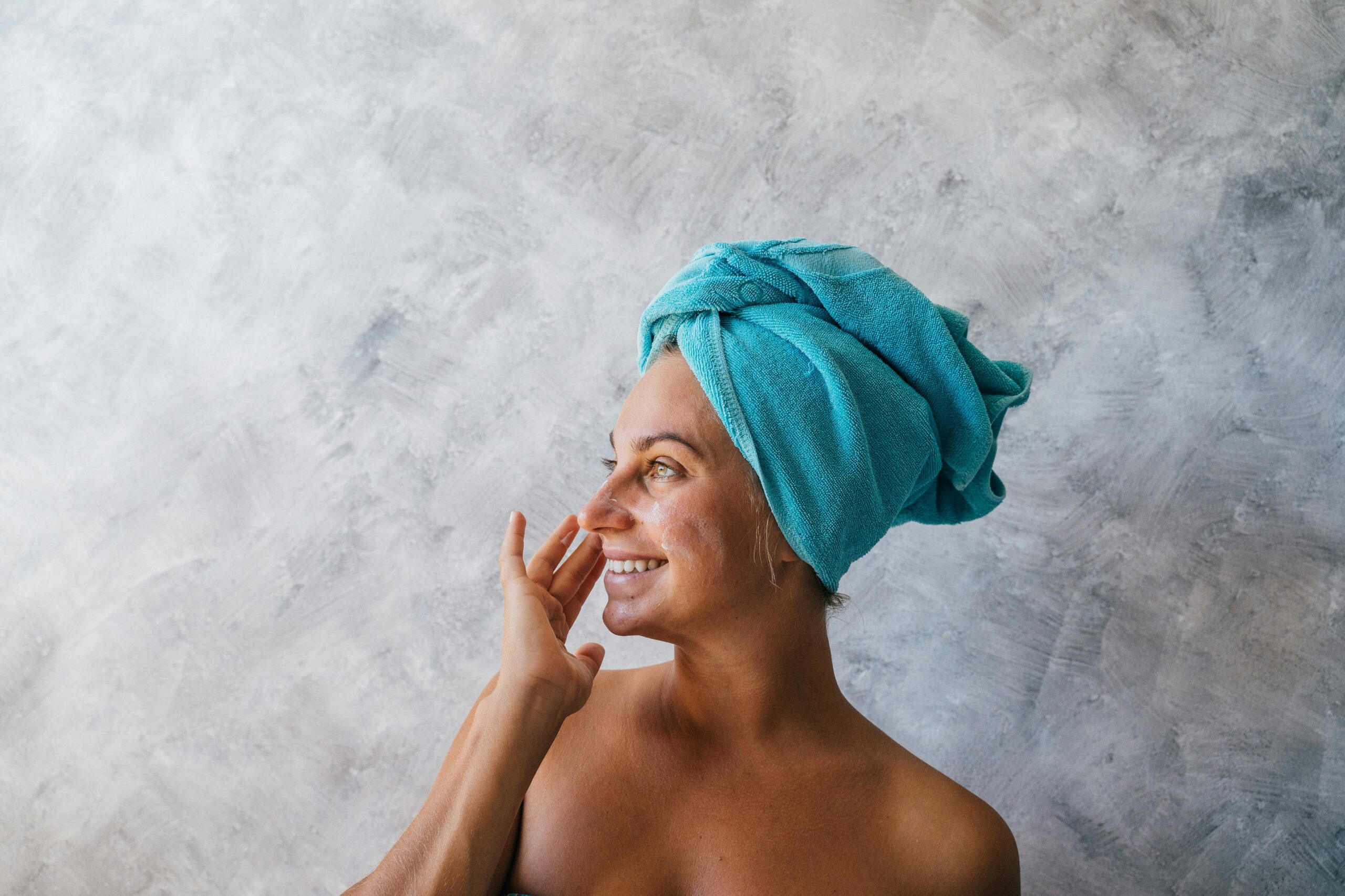 Smiling woman applying facial cream with a towel wrapped on her head.